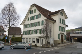 Seitenansicht mit Auto im Vordergrund, gelbe Fassade und braunem Dach. 